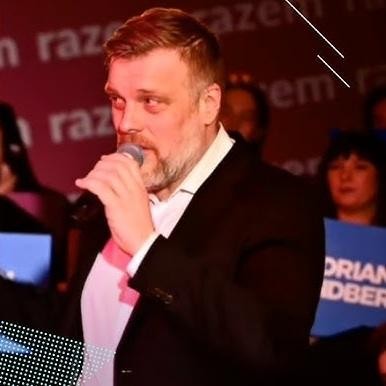 Adrian Zandberg przemawiający podczas wystąpienia publicznego, z widocznym zaangażowaniem i gestykulacją.