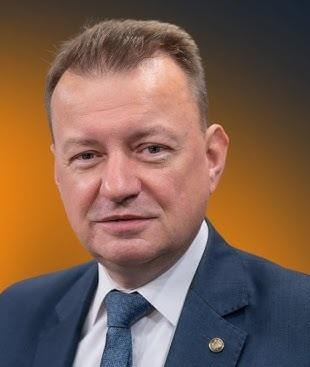 Mariusz Błaszczak przemawiający na wiecu politycznym, z energicznym gestem i zdeterminowanym wyrazem twarzy, na tle niewyraźnego tłumu.