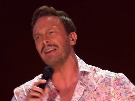 Sławek Uniatowski na scenie podczas występu, otoczony dynamicznym oświetleniem, z elementami instrumentów muzycznych w tle, ukazujący jego sceniczną energię i pasję.
