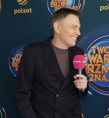 Krzysztof Ibisz podczas prowadzenia teleturnieju, w dynamicznej pozie na tle kolorowego studia telewizyjnego, z uśmiechem na twarzy.